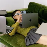 Una chica con gafas de sol, recostada en un sofá verde entre varios portátiles y su móvil